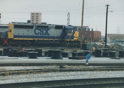 A side view of a CSX diesel locomotive, number 8247, parked in a gravel rail yard with power lines, buildings, and parked cars in the background under an overcast sky.