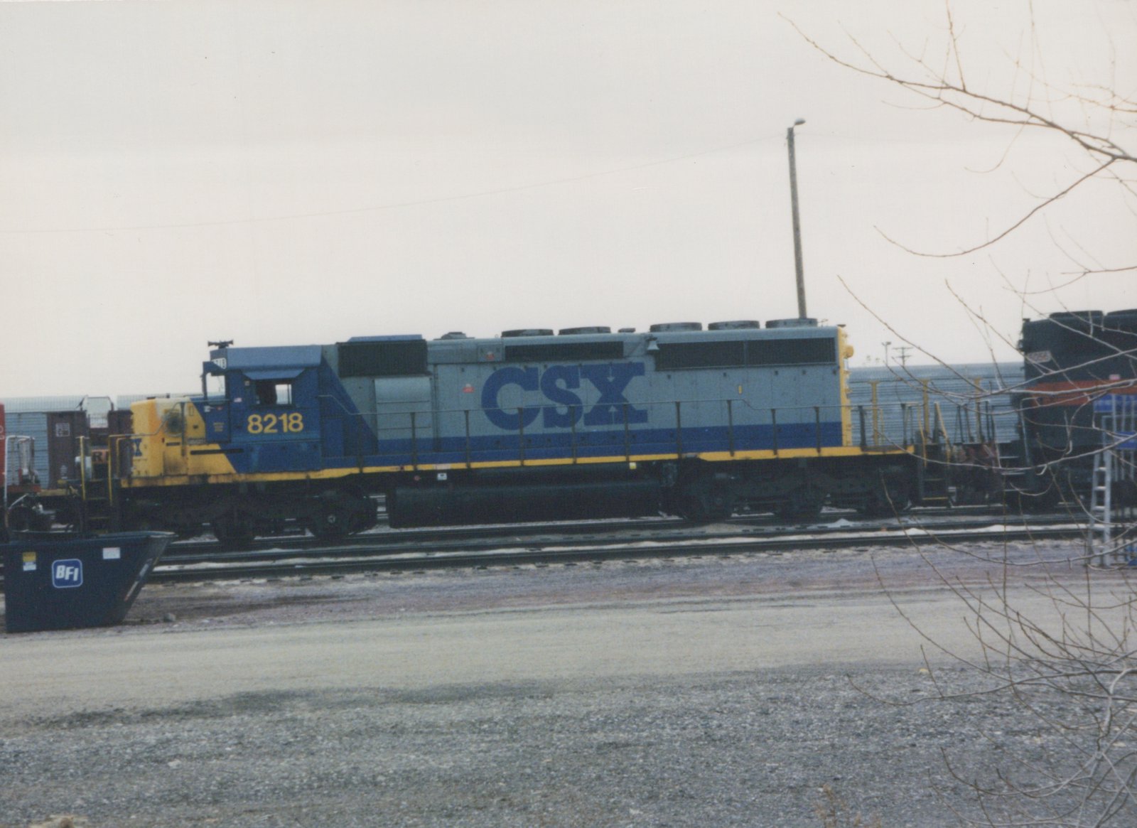 A side view of a gray, blue, and yellow CSX diesel locomotive numbered 8218, standing on tracks in a rail yard on an overcast winter day, with a blue dumpster in the foreground.