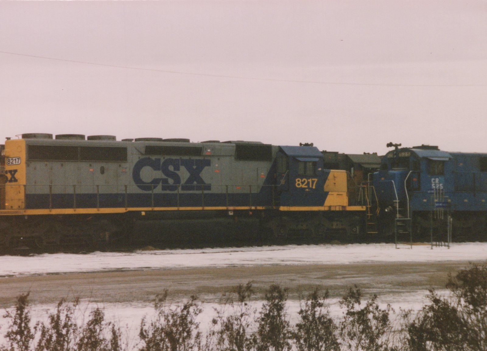 Two CSX diesel locomotives, one numbered 8217 and the other CR 525, are parked side by side in a snowy yard under an overcast sky.