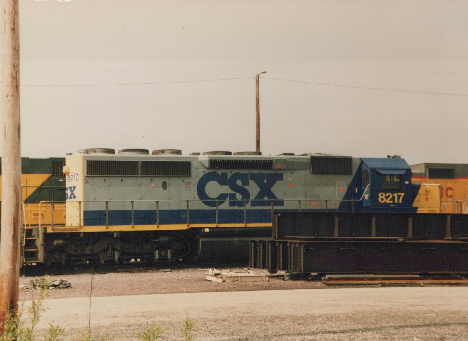 Side view of a blue, gray, and yellow CSX diesel locomotive, number 8217, parked on tracks in a gravel yard under an overcast sky.