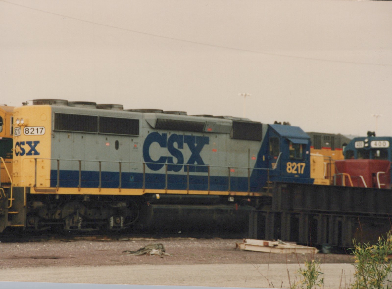 A side view of a gray, blue, and yellow CSX diesel locomotive, number 8217, in a gravel yard with other trains in the background.
