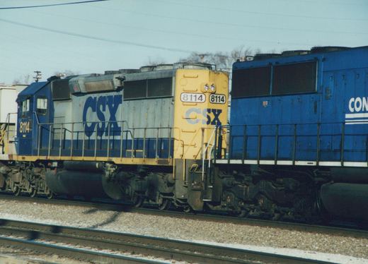 A side view of a CSX locomotive numbered 8114, with a blue and white body and yellow front, on train tracks with a second blue locomotive behind it.