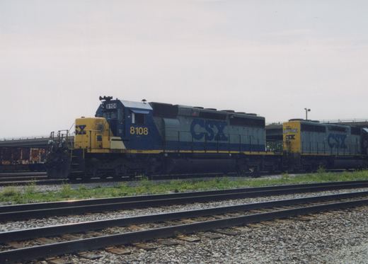 Two blue and gray CSX locomotives with yellow fronts are parked on adjacent tracks in a gravel yard with weeds growing between the rails; the lead unit is numbered 8108 and the trailing unit is 8430.