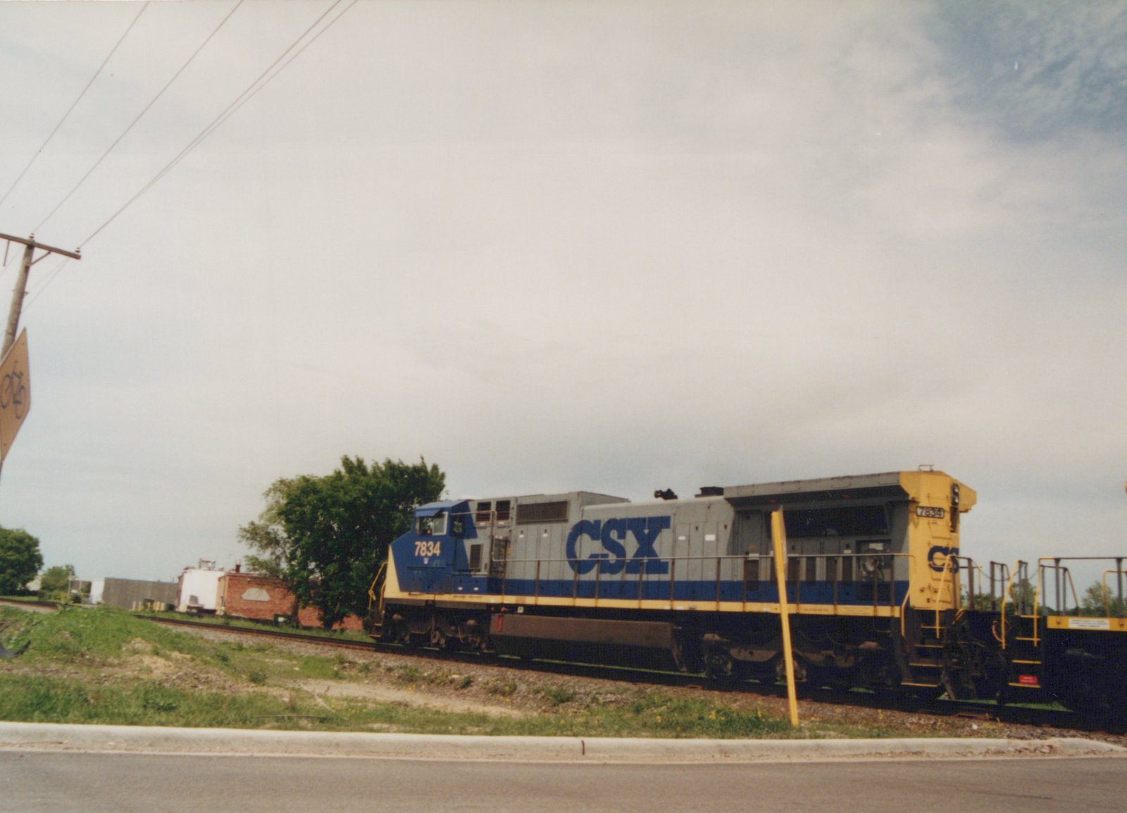 A CSX Transportation diesel locomotive numbered 7834, with a gray, blue, and yellow livery, is on a railroad track next to a grassy embankment and a street under a cloudy sky.