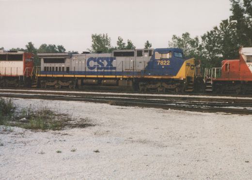 A side view of a blue and yellow CSX GP38-2 locomotive, number 7822, standing on tracks in a gravel yard, with a red and white CSX SD40-2, number 6010, visible behind it.