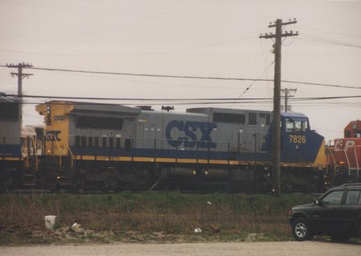 A side view of a gray, blue, and yellow CSX diesel locomotive numbered 7826 on a mainline track, with a red Canadian Pacific locomotive behind it and a black SUV parked in the foreground.