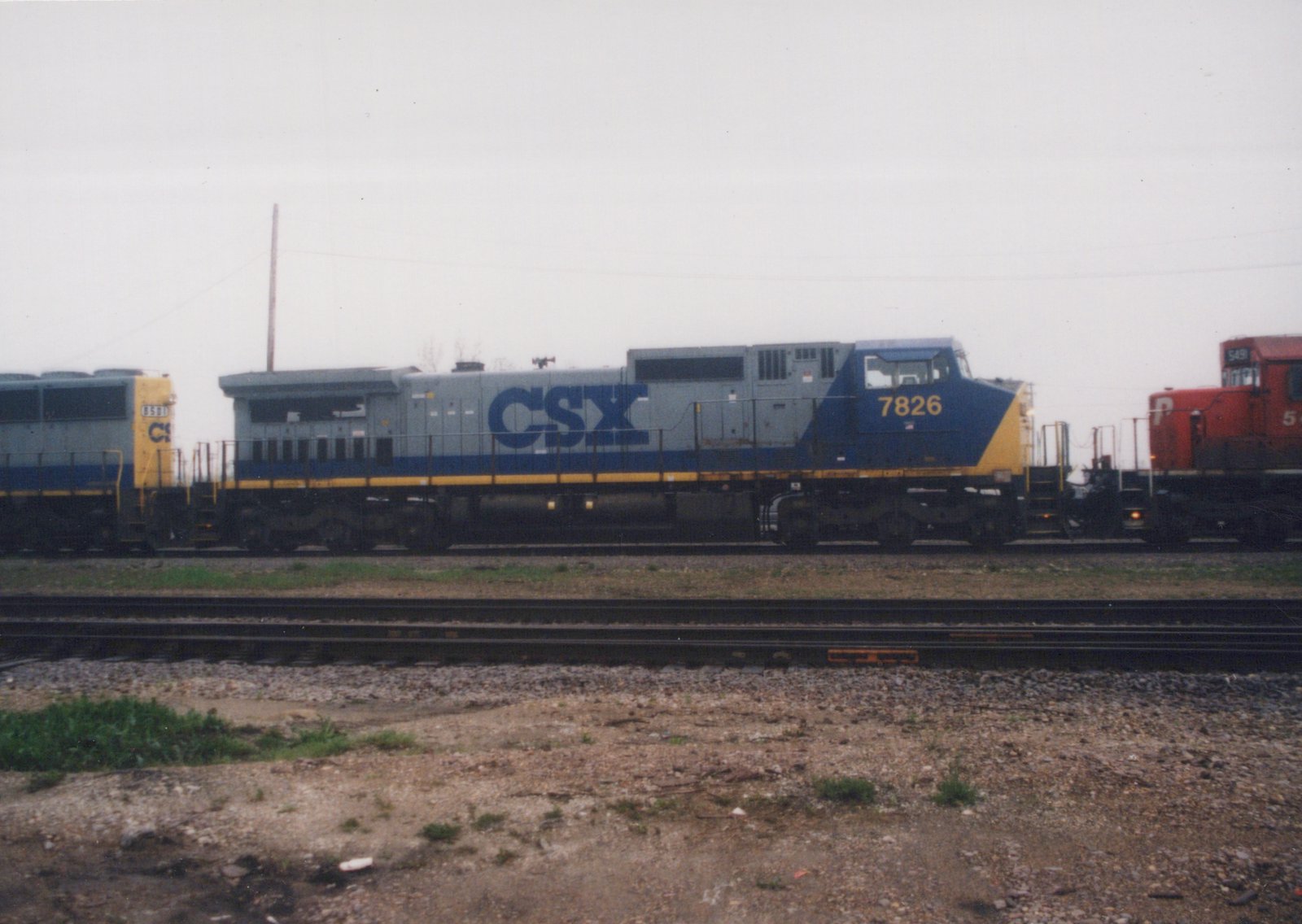 A side view of a CSX locomotive with number 7826 on its front, coupled to another CSX unit 6591, with a red Penn Central locomotive 549 in the background, all on tracks in a gravel yard under an overcast sky.