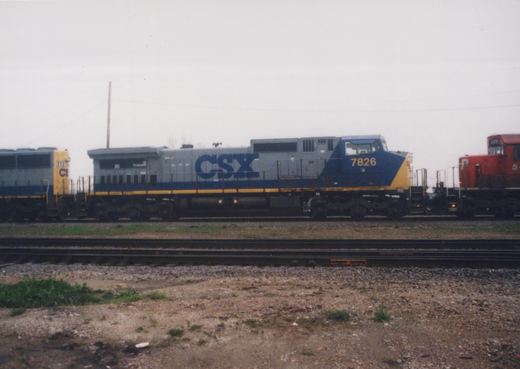 A side view of a CSX locomotive with number 7826 on its front, coupled to another CSX unit 6591, with a red Penn Central locomotive 549 in the background, all on tracks in a gravel yard under an overcast sky.