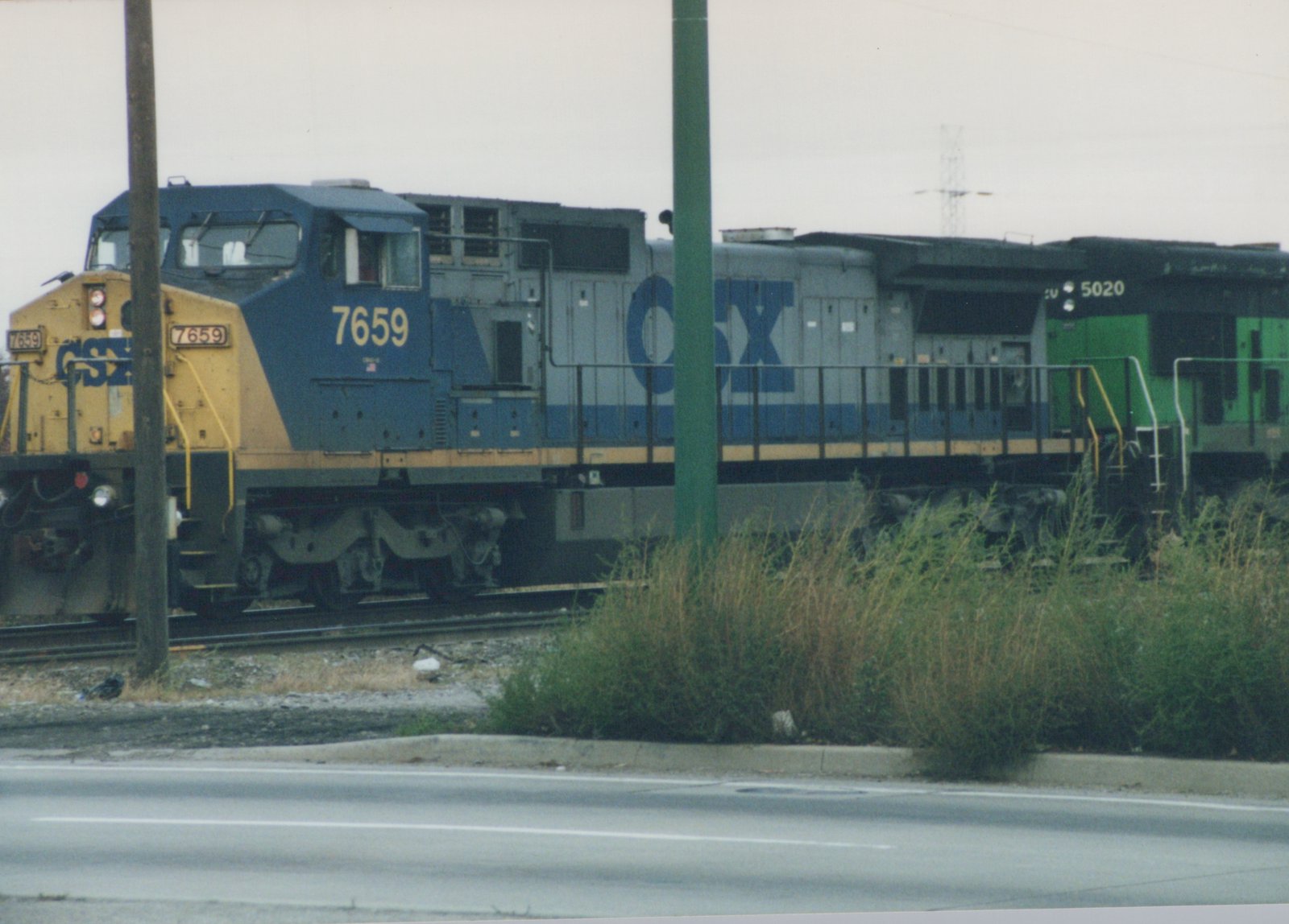 A blue and yellow CSX locomotive numbered 7659 is shown on the tracks from a three-quarter front view, with a second green locomotive behind it, viewed from across a road with overgrown weeds.