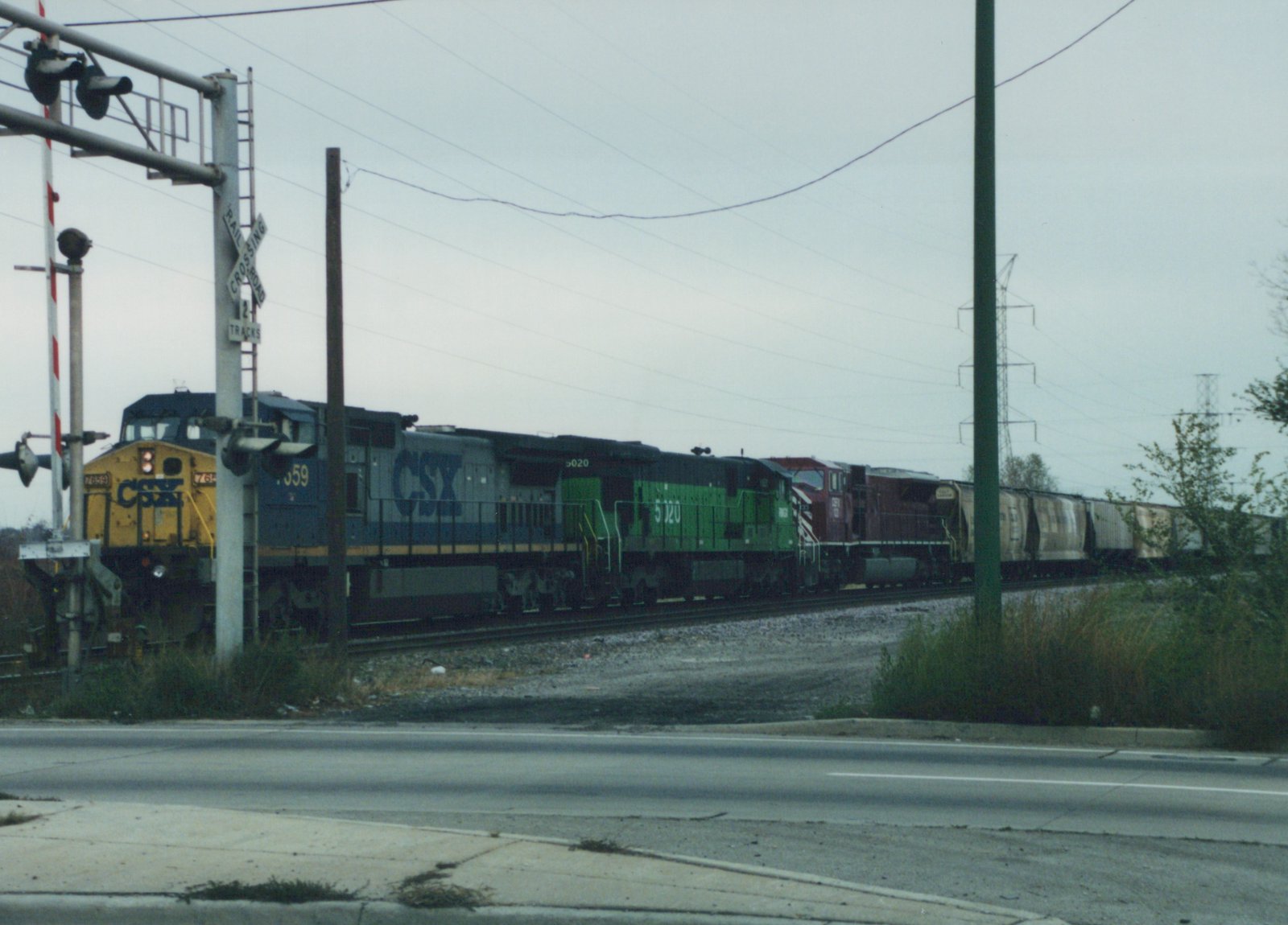 A CSX freight train led by a yellow and blue GP38-2 and a green and black SD40-2 locomotive passes a railroad crossing on an overcast day.