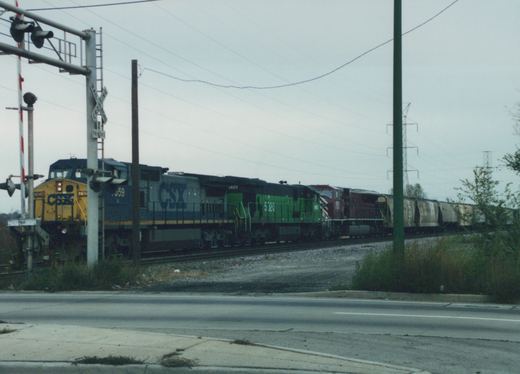 A CSX freight train led by a yellow and blue GP38-2 and a green and black SD40-2 locomotive passes a railroad crossing on an overcast day.