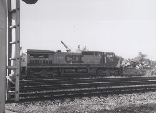 A black and white photo of a CSX diesel locomotive, number 7712, on a set of tracks with a mobile crane in the background. The locomotive is shown from the side.