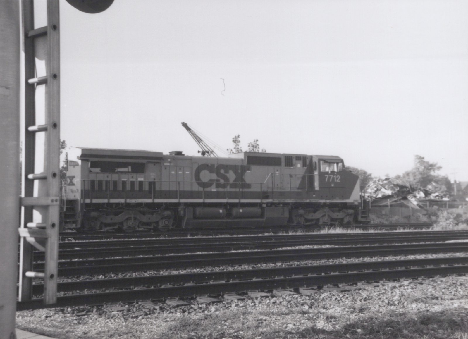 A black and white photo of a CSX diesel locomotive, number 7712, on a set of tracks with a mobile crane in the background. The locomotive is shown from the side.