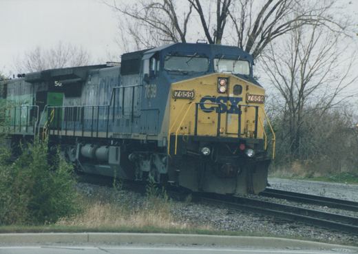 A blue and yellow CSX diesel locomotive numbered 7659 is moving on train tracks next to a road, with leafless trees in the background.