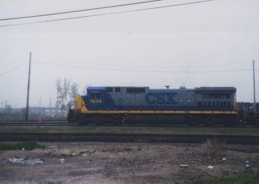 Side view of a blue and yellow CSX diesel locomotive, number 7644, on multiple tracks in a gravel yard under a gray sky.