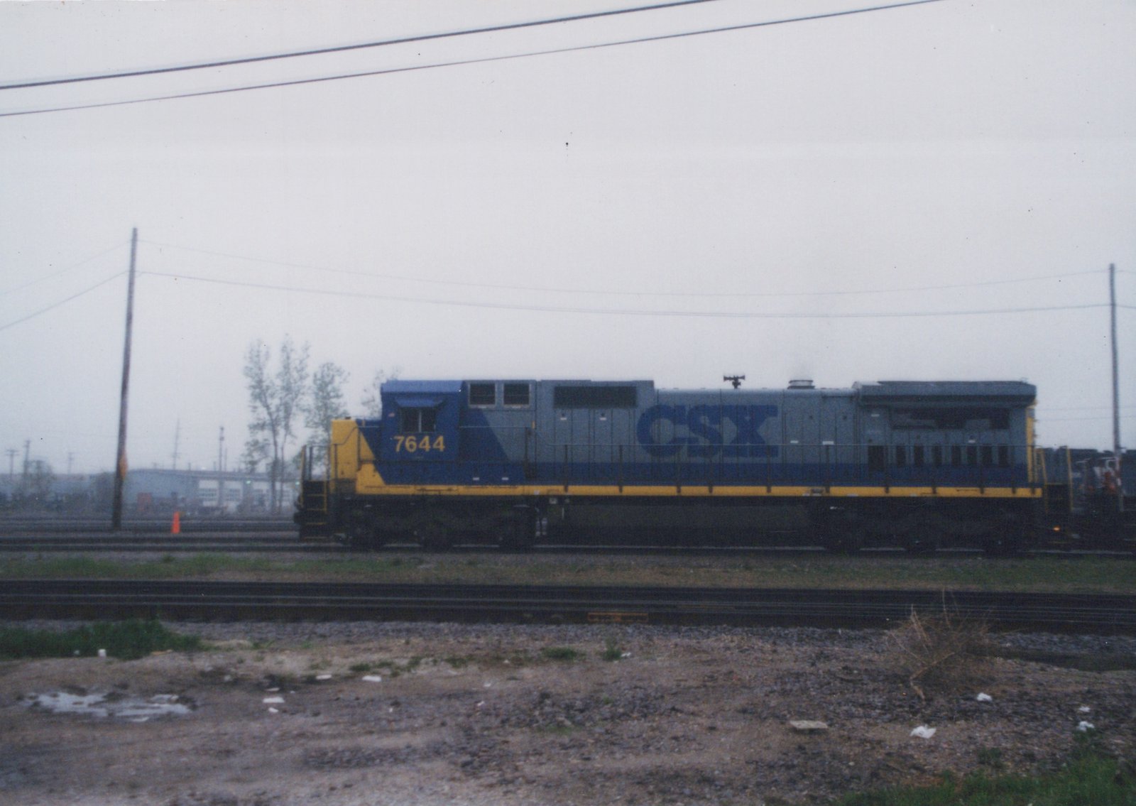 Side view of a blue and yellow CSX diesel locomotive, number 7644, on multiple tracks in a gravel yard under a gray sky.
