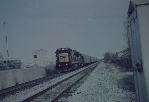 A blue and yellow CSX locomotive numbered 7644 is seen pulling a long freight train on a snowy, overcast day along a track beside a concrete wall and buildings.