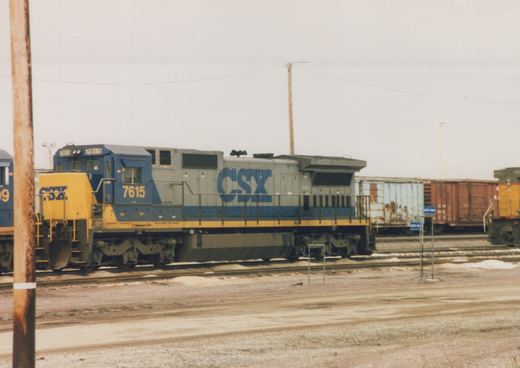 A blue and yellow CSX diesel locomotive with the number 7615 is parked on a set of tracks in a railyard. Snow is visible on the ground, and freight cars are in the background.