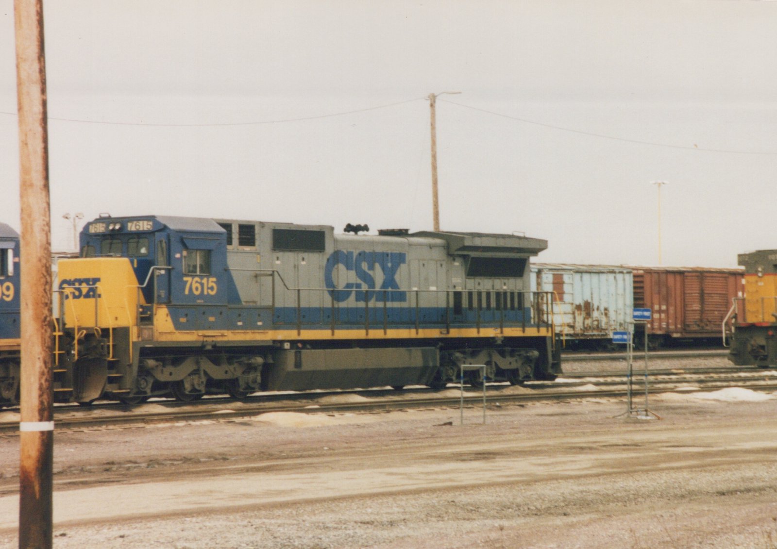 A blue and yellow CSX diesel locomotive with the number 7615 is parked on a set of tracks in a railyard. Snow is visible on the ground, and freight cars are in the background.