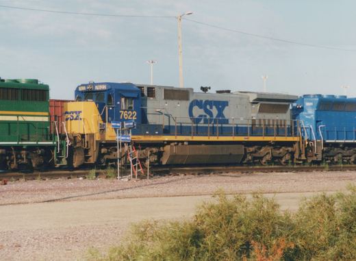 Side view of a blue, gray, and yellow CSX locomotive numbered 7622 in a gravel rail yard under a clear sky, with another blue locomotive numbered 3131 behind it.