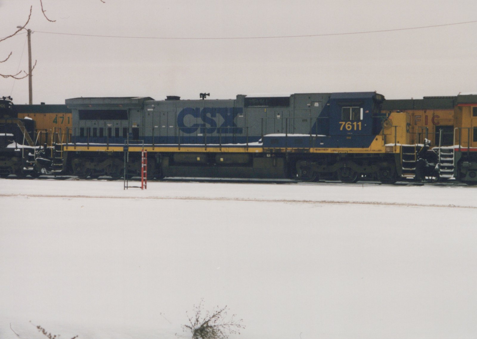 A side view of a gray, blue, and yellow CSX diesel locomotive, number 7611, parked on tracks in a snowy yard with other locomotives nearby.