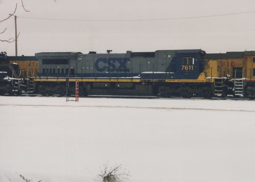 A side view of a gray, blue, and yellow CSX diesel locomotive, number 7611, parked on tracks in a snowy yard with other locomotives nearby.