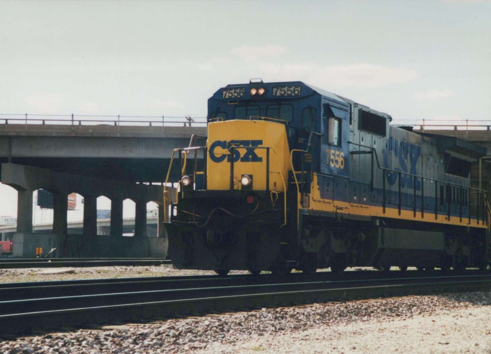 A blue and yellow CSX diesel locomotive, number 7556, sits on railroad tracks beneath a large concrete overpass on a sunny day.