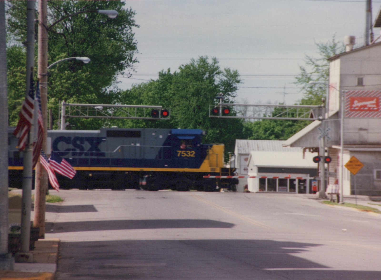 A blue and yellow CSX diesel locomotive with the number 7532 is stopped at a grade crossing with flashing red lights and lowered gates, surrounded by trees and buildings.
