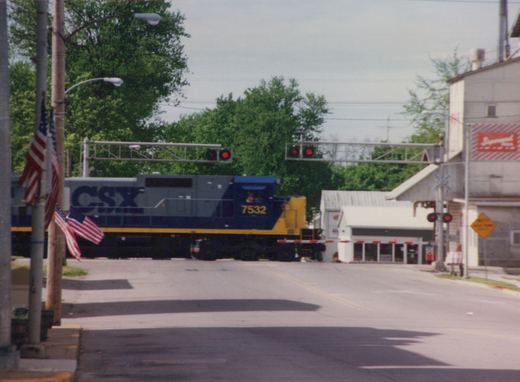 A blue and yellow CSX diesel locomotive with the number 7532 is stopped at a grade crossing with flashing red lights and lowered gates, surrounded by trees and buildings.