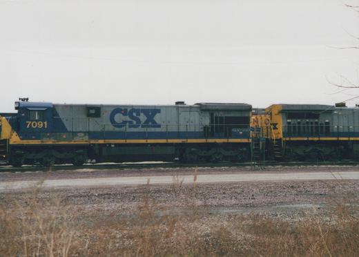 A side view of two CSX diesel locomotives, numbered 7091 and 368, parked on a track in a gravel yard with dry grass in the foreground.