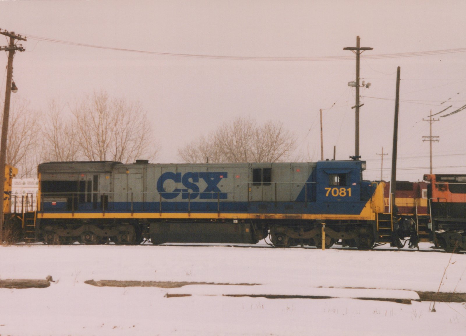 A side view of a CSX diesel locomotive numbered 7081, parked on tracks in a snowy yard with another locomotive behind it.
