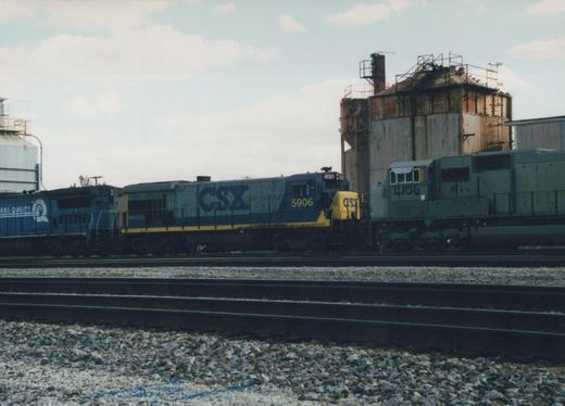 A blue and yellow CSX locomotive numbered 5906 is parked on multiple tracks in a yard, with a gray locomotive numbered 4106 beside it and industrial buildings in the background.