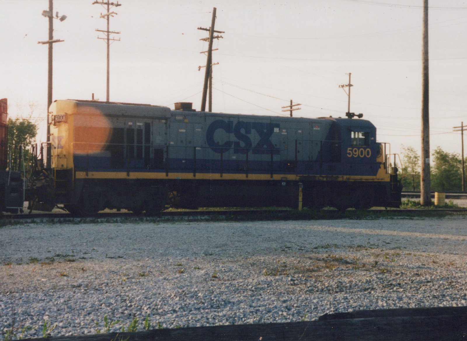 A blue and yellow CSX diesel locomotive numbered 5900 sits on railroad tracks in a gravel yard with utility poles in the background on a clear day.