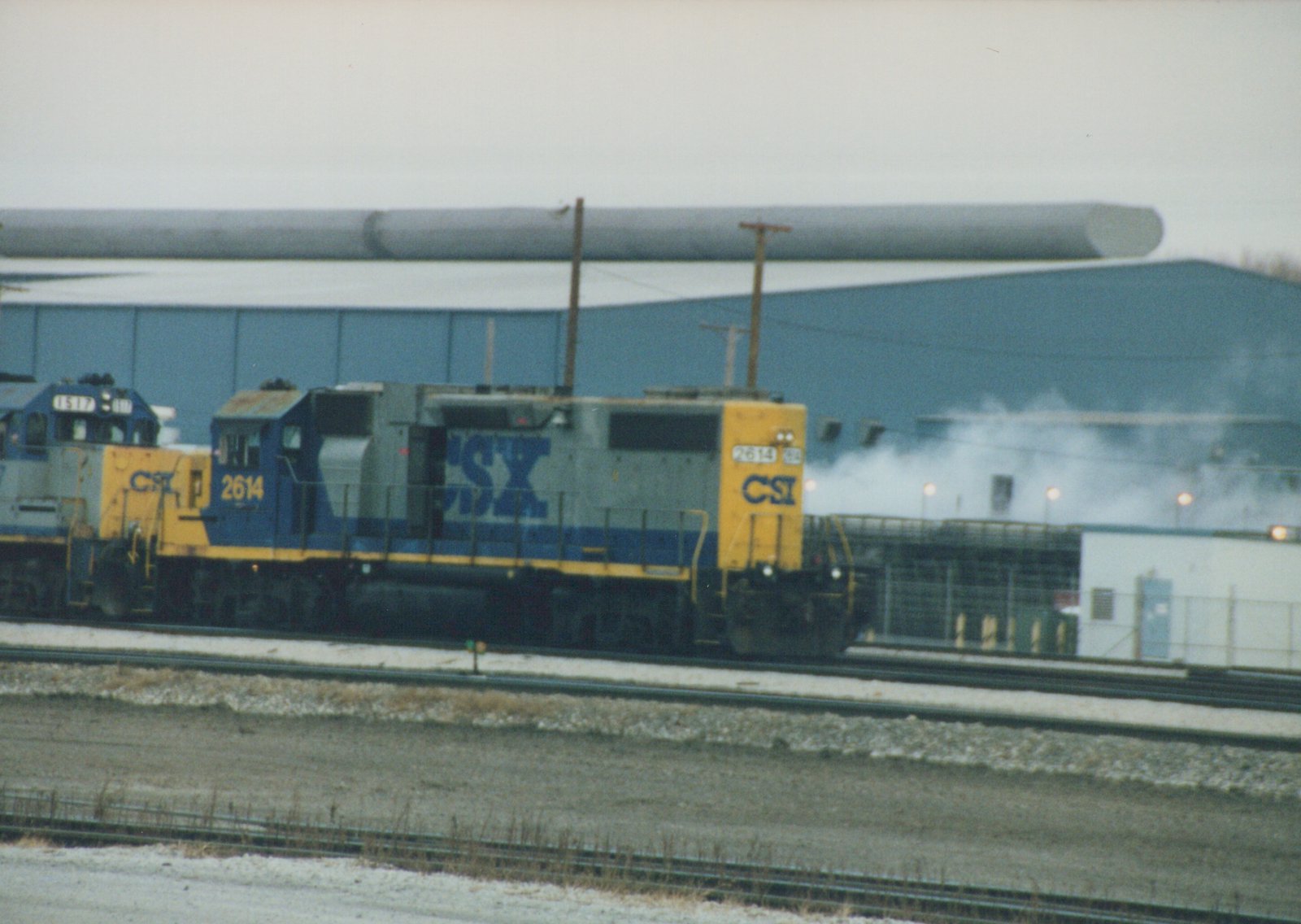 A blue and yellow CSX diesel locomotive, number 2614, is shown on the tracks in front of a large industrial building with snow on the ground, followed by another locomotive, number 1517.