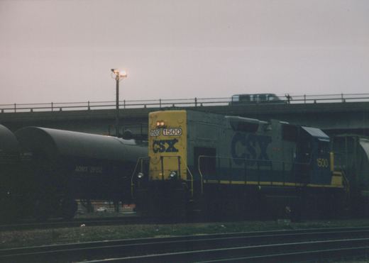 A yellow and blue CSX diesel locomotive numbered 1500 is shown front and side, pulling tank cars under a concrete overpass on a cloudy day.