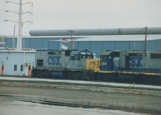 Two CSX diesel locomotives, numbered 1517 and 2614, sit side by side on a snowy railroad yard in front of a large industrial building under an overcast sky.