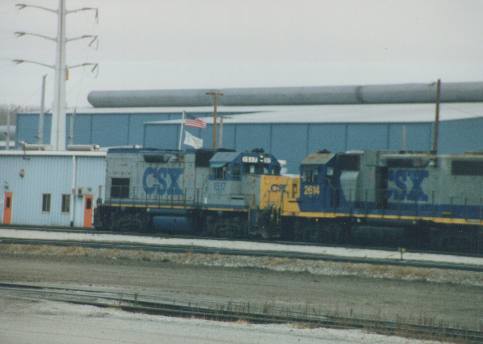 Two CSX diesel locomotives, numbered 1517 and 2614, sit side by side on a snowy railroad yard in front of a large industrial building under an overcast sky.