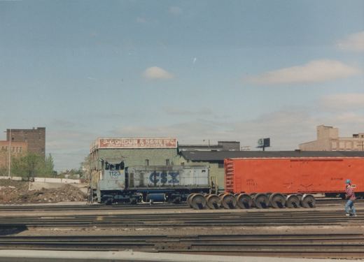 A white and blue CSX locomotive with the number 1123 is coupled to a bright orange Amtrak boxcar in a railway yard. A person in a blue hard hat walks on the tracks nearby, with industrial buildings and a clear sky in the background.