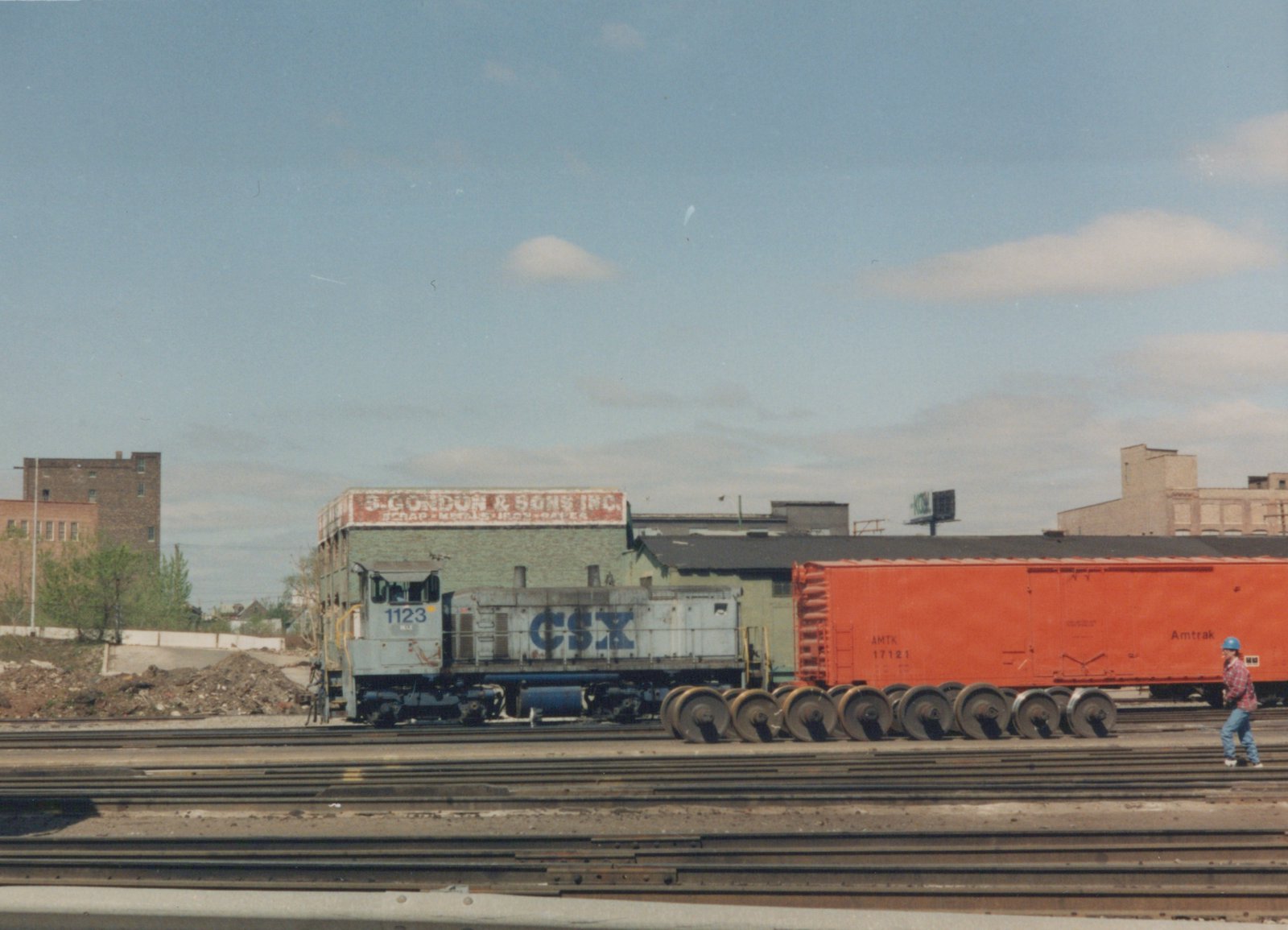 A white and blue CSX locomotive with the number 1123 is coupled to a bright orange Amtrak boxcar in a railway yard. A person in a blue hard hat walks on the tracks nearby, with industrial buildings and a clear sky in the background.