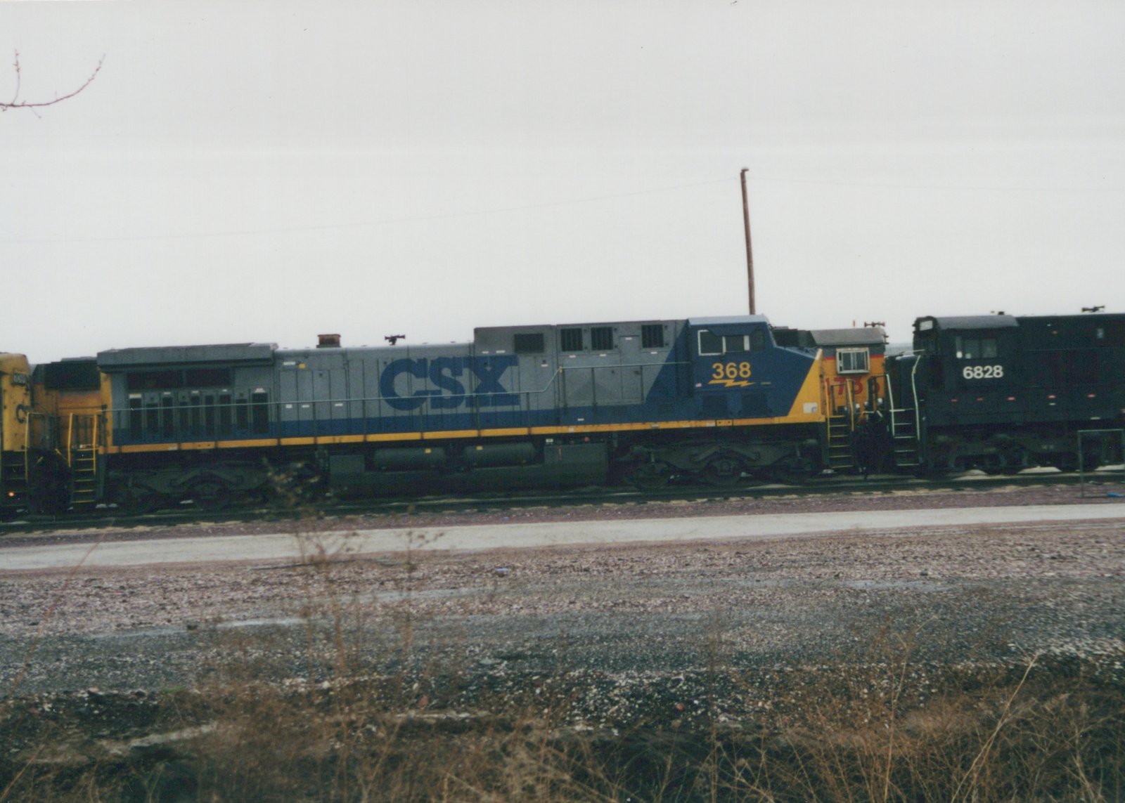 Side view of a gray CSX freight train with blue and yellow stripes, featuring locomotive 368 in the front and 6828 behind it, on a railroad main line in a yard with bare ground and overcast sky.