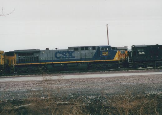Side view of a gray CSX freight train with blue and yellow stripes, featuring locomotive 368 in the front and 6828 behind it, on a railroad main line in a yard with bare ground and overcast sky.