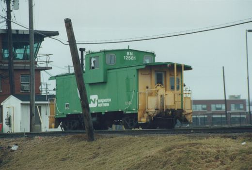 A green and yellow Burlington Northern caboose numbered 12581 is parked on train tracks beside a brick building, with a grassy slope in the foreground under an overcast sky.