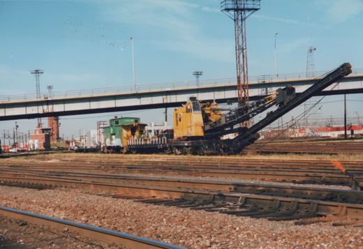 A yellow track maintenance machine and a green and yellow locomotive sit on a series of railroad tracks in a railyard under a clear blue sky, with an elevated highway bridge in the background.