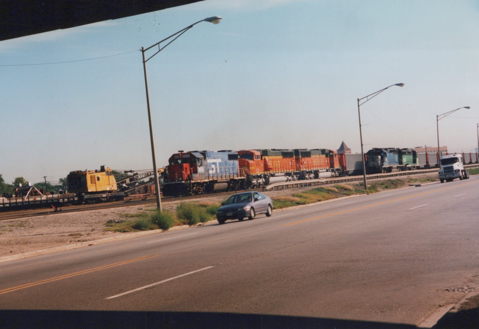 A photo of three diesel locomotives from BNSF and Norfolk Southern on a rail line next to a street with a silver sedan and a white semi-truck, taken in Cicero, Illinois.