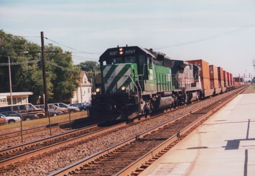 A green and white Burlington Northern diesel locomotive, number 8087, pulls a long freight train of container cars past a station platform. Parked cars and green trees are visible in the background under a clear sky.