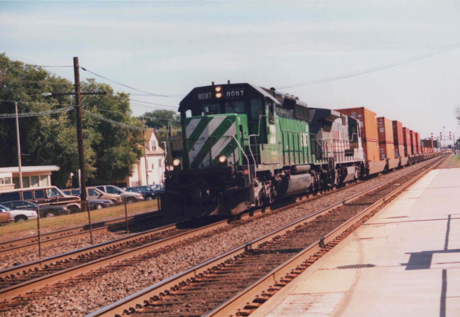 A green and white Burlington Northern diesel locomotive, number 8087, pulls a long freight train of container cars past a station platform. Parked cars and green trees are visible in the background under a clear sky.