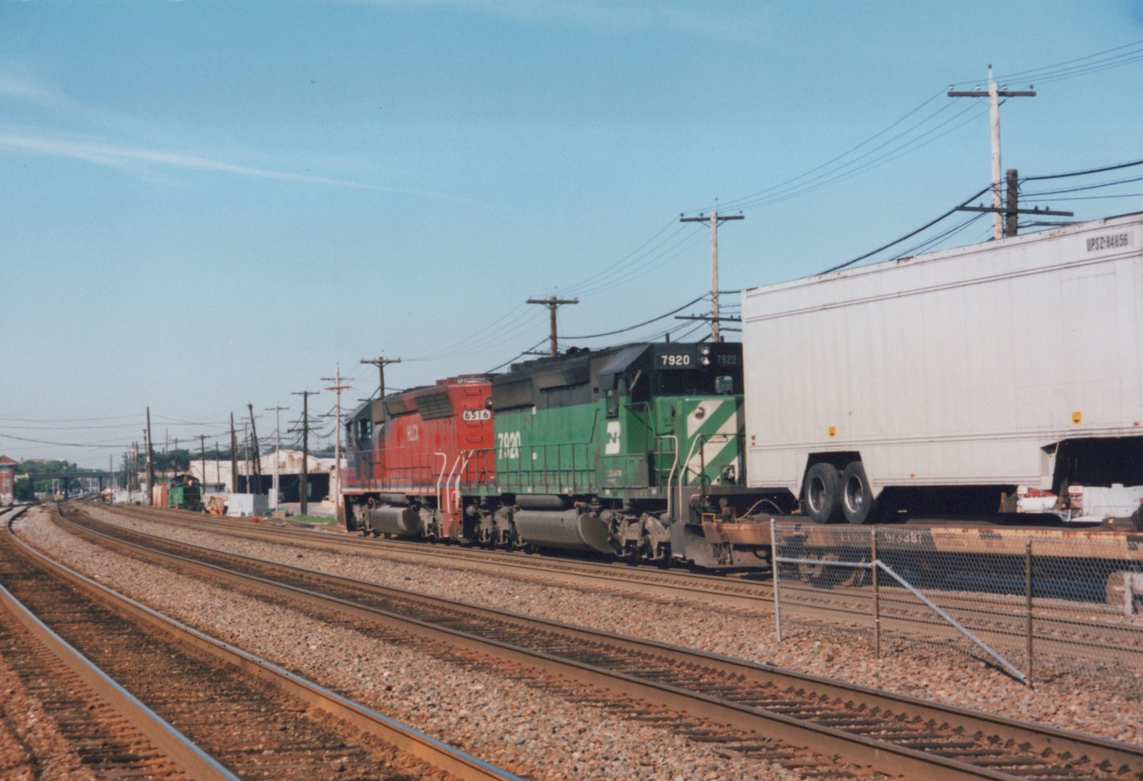 Two diesel locomotives, one green and one red, pull a freight train with a white semi-trailer on a flatcar along a railroad main line under a clear sky.