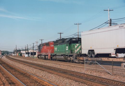 Two diesel locomotives, one green and one red, pull a freight train with a white semi-trailer on a flatcar along a railroad main line under a clear sky.