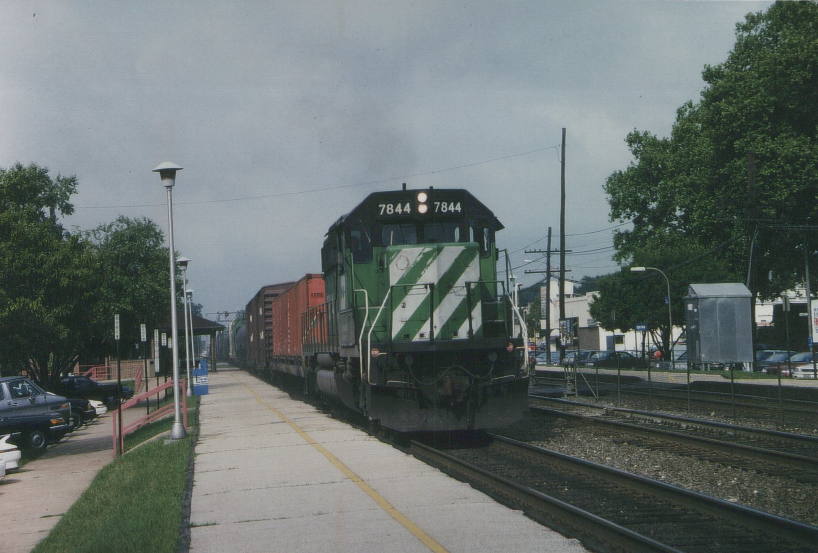 A green and white striped Burlington Northern diesel locomotive numbered 7844 is stopped at a station platform, pulling a line of freight cars under an overcast sky.