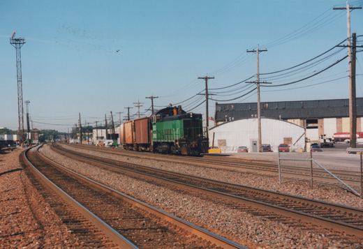 A green and black diesel locomotive with the letters BNSF on its side is moving along a set of railroad tracks, pulling several boxcars. The background shows industrial buildings and a clear blue sky. Several cars are parked nearby.